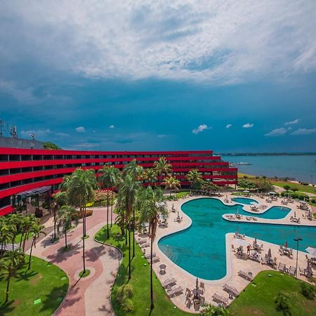 Royal Tulip Brasilia Alvorada Hotel Exterior photo The photo shows a modern hotel building with a striking red exterior, surrounded by lush greenery. In the foreground, there is a large, creatively shaped swimming pool, with lounge chairs arranged around it. The backdrop features a body of water, pos